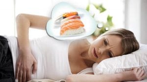 blond woman in white t-shirt, doubled over in pain, lying in bed, with thought bubble displaying salmon fillet